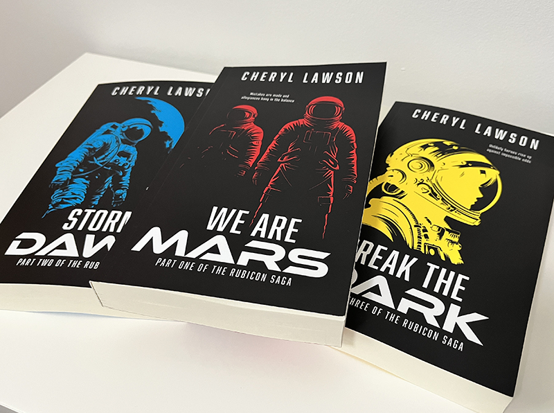The Rubicon Saga books - We Are Mars, Storm At Dawn, Break the Dark covers with astronaut illustrations. Book covers shown against a blue illustrated space ship interior.